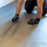 A professional cleaner cleaning grout with a brush blade and foamy soap on a gray tiled bathroom floor